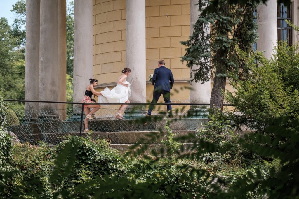 Hochzeitskrone Einer Braut wird beim Anziehen ihres Kleides geholfen, während sie und ein Mann vor einem von Grün umgebenen Gebäude eine Treppe hinaufsteigen. Ein Hochzeitsfotograf in Wien und Niederösterreich hielt die Szene fest und schuf so authentische Erinnerungen an den besonderen Tag.