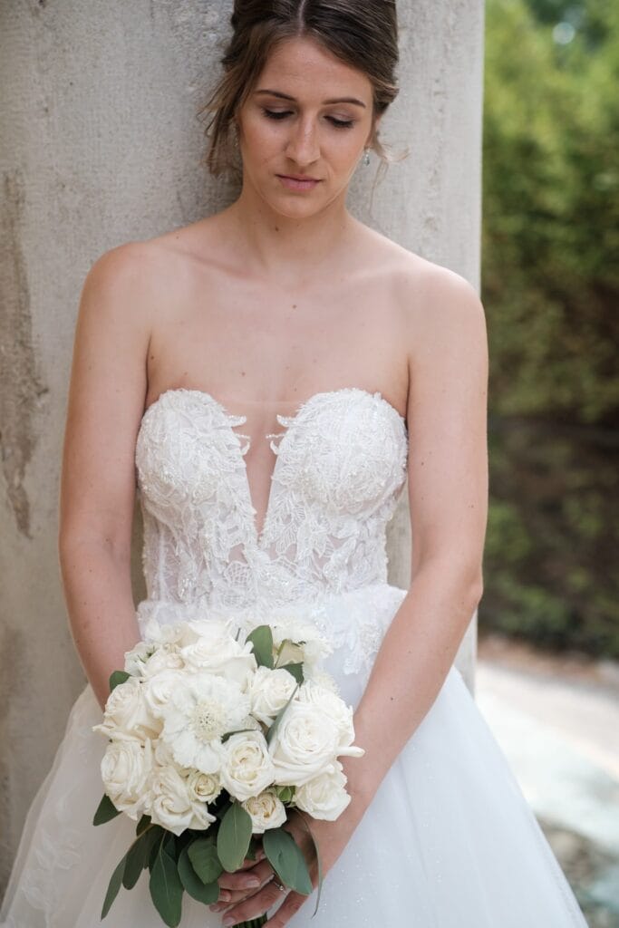 Hochzeitskrone Braut in einem weißen Spitzenhochzeitskleid mit einem Strauß weißer Rosen, steht im Freien vor einer Steinmauer. Von einem Hochzeitsfotografen in Wien und Niederösterreich festgehalten, fühlt sich jeder Moment echt und elegant an, perfekt für alle, die natürliche Hochzeitsfotos suchen.