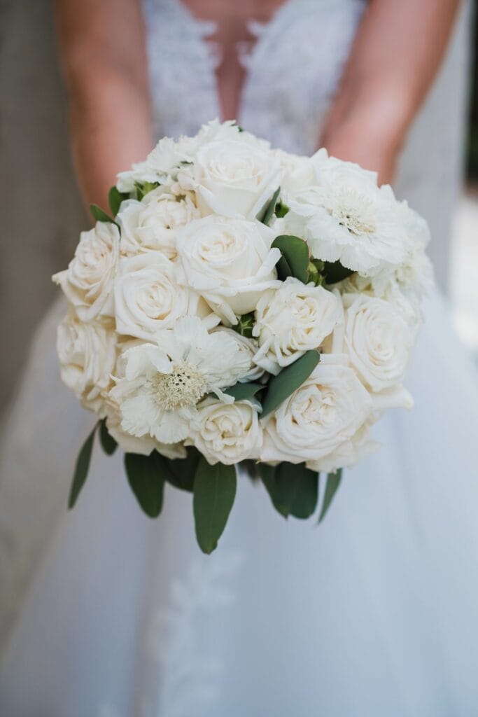 Hochzeitskrone Eine Braut hält einen Strauß weißer Rosen und Grünpflanzen, aufgenommen von einem Hochzeitsfotografen in Wien und Niederösterreich, wobei ihr Hochzeitskleid im Hintergrund kunstvoll unscharf gemacht ist.