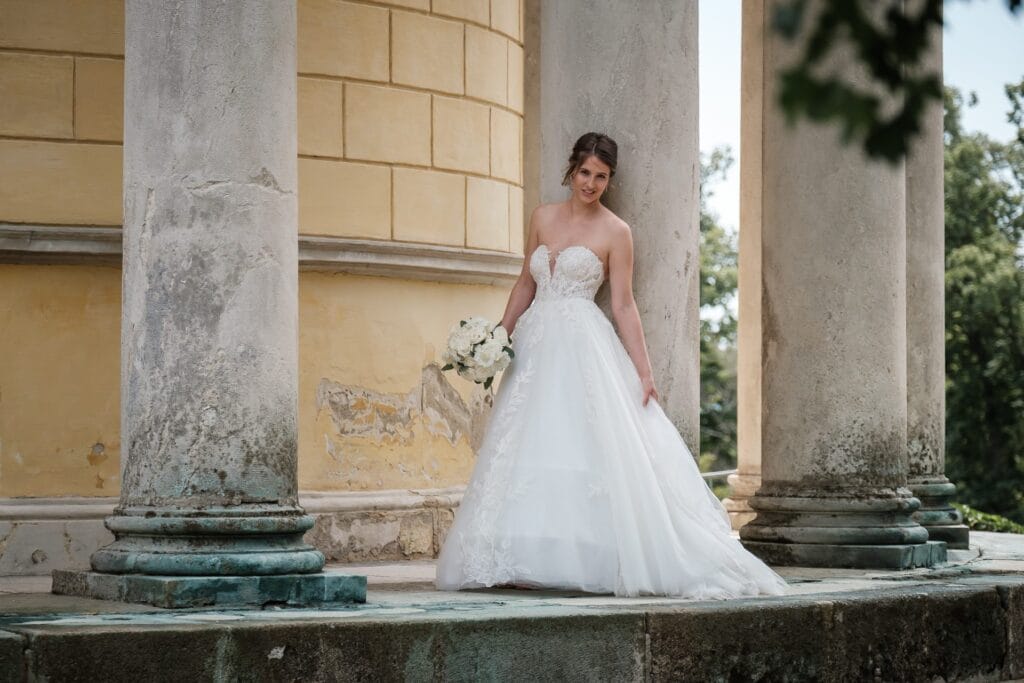 Hochzeitskrone Eine Braut in einem weißen Brautkleid steht zwischen Steinsäulen und hält einen Strauß weißer Blumen in der Hand. Dieser Hochzeitsfotograf in Wien und Niederösterreich fängt authentische Momente ein und kreiert natürliche Hochzeitsfotos, die die Essenz Ihres besonderen Tages für immer festhalten.