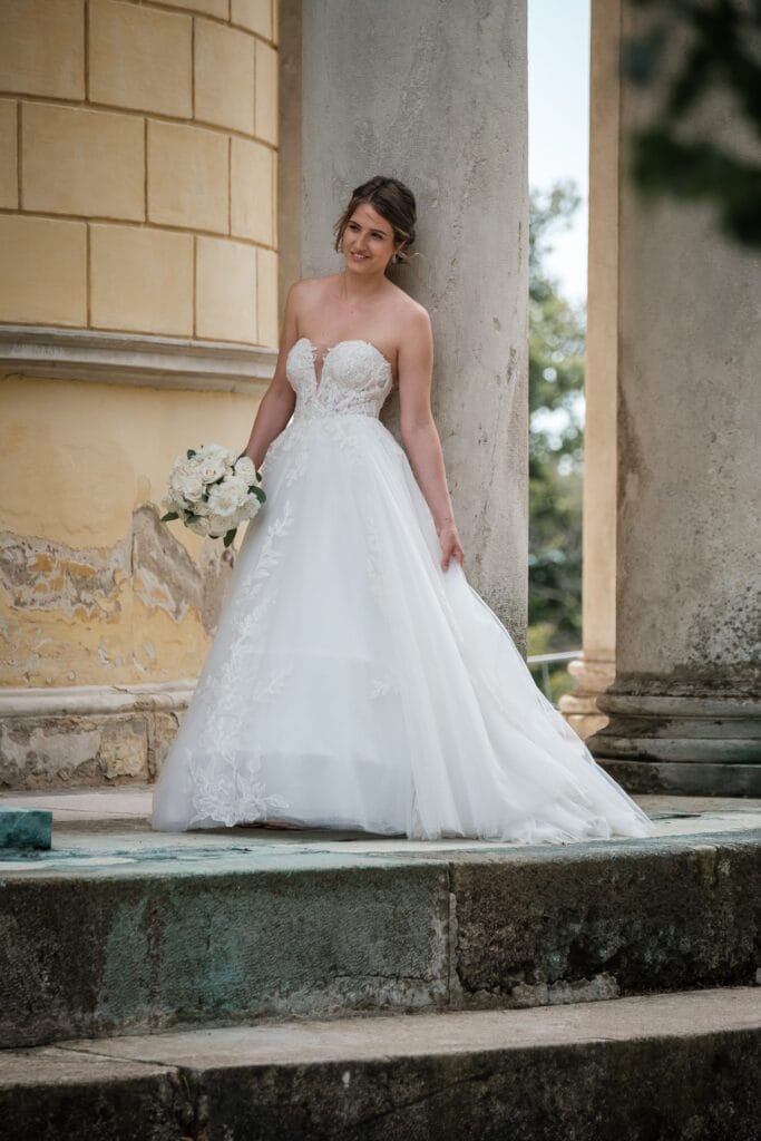 Hochzeitskrone Eine Braut in einem trägerlosen weißen Kleid mit einem Blumenstrauß steht auf Steinstufen in der Nähe von Säulen und einem gelben Gebäude, aufgenommen von einem Hochzeitsfotografen in Wien und Niederösterreich, wodurch authentische Hochzeitsfotos entstehen.