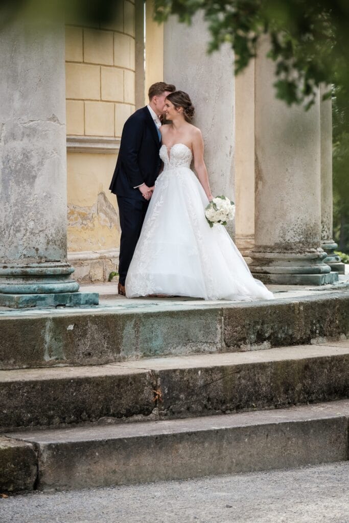 Hochzeitskrone Zwischen Steinsäulen küsst sich ein Paar im Hochzeitskleid zärtlich. Die Braut umklammert ihren Blumenstrauß, während der Bräutigam im dunklen Anzug ihre Hand hält. Ihre Umarmung fängt authentische Hochzeitsfotos ein, die wahre Momente der Liebe und Verbundenheit wunderschön festhalten.