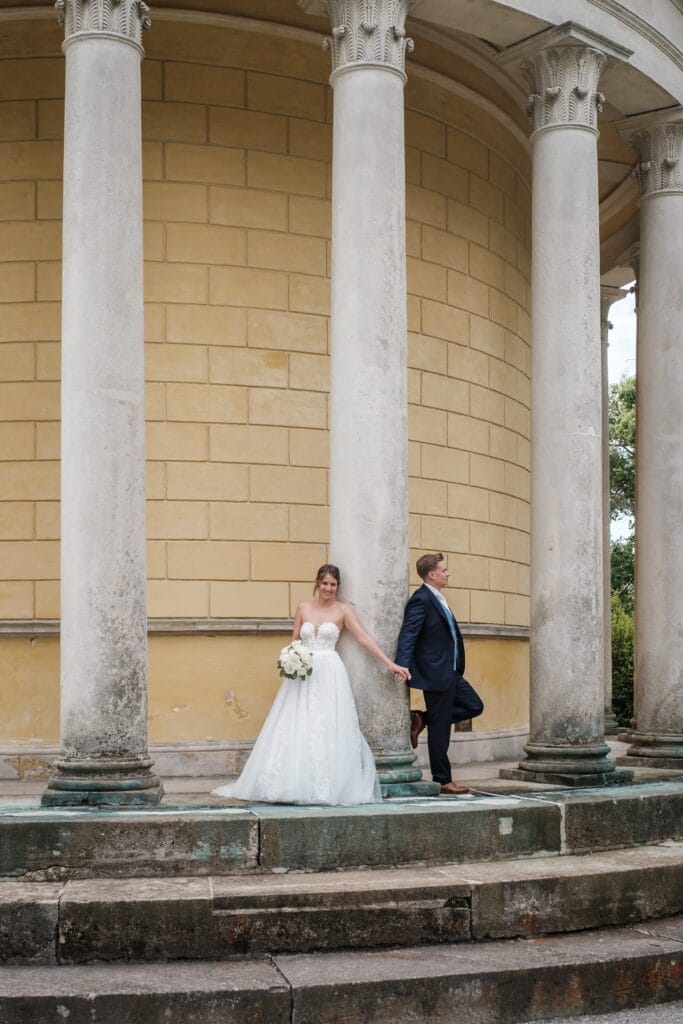 Hochzeitskrone Braut und Bräutigam stehen auf Steinstufen, durch eine Säule getrennt, festgehalten in authentischen Hochzeitsfotos. Sie hält einen Blumenstrauß in ihrem weißen Kleid, während er einen dunklen Anzug trägt; beide blicken in entgegengesetzte Richtungen.