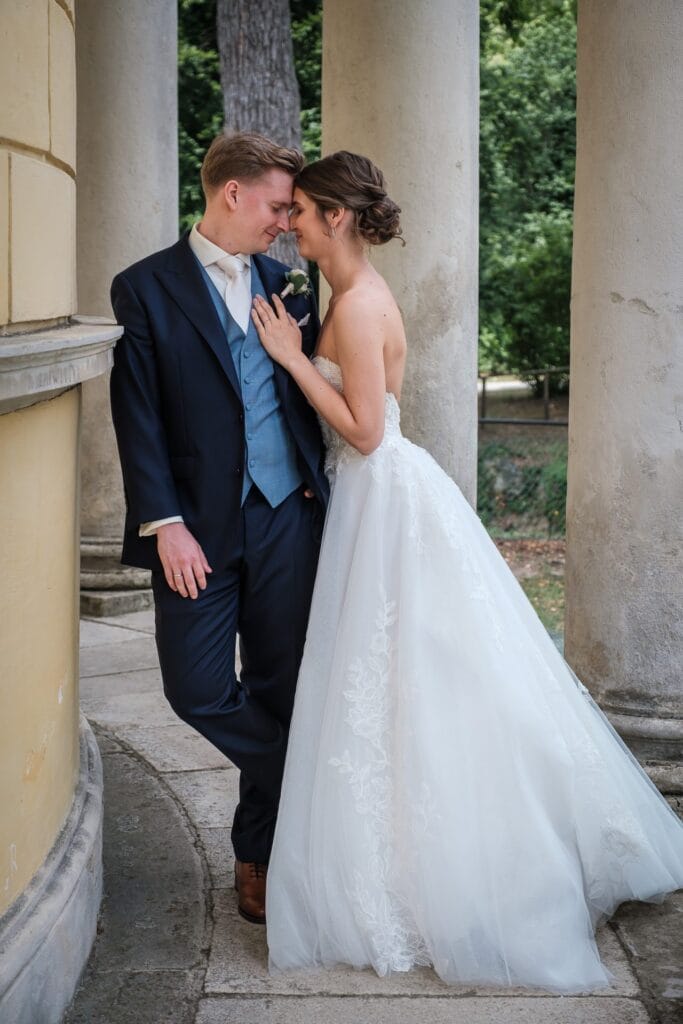 Hochzeitskrone Eine Braut im weißen Kleid und ein Bräutigam im dunklen Anzug stehen dicht beieinander und lehnen an einer Steinkonstruktion mit Säulen. Sie stehen einander gegenüber und berühren ihre Köpfe. Ein Hochzeitsfotograf in Wien und Niederösterreich hat sie in echten Momenten festgehalten.