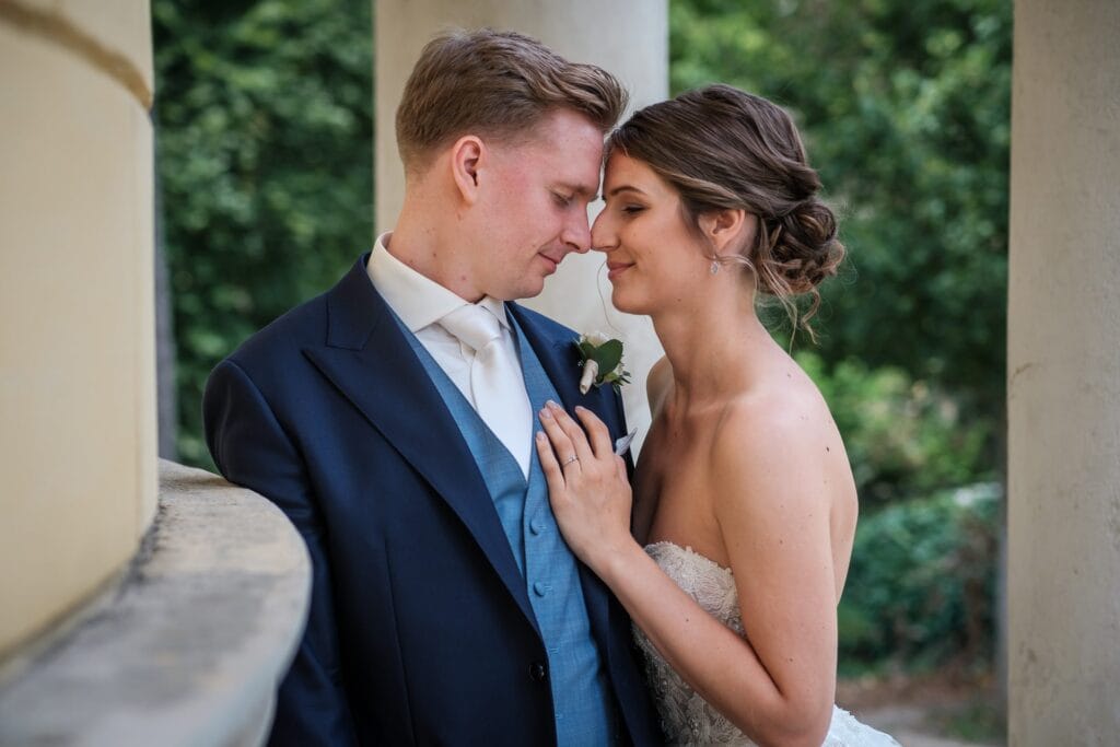 Hochzeitskrone Ein Paar im Hochzeitskleid teilt einen intimen Moment und berührt seine Stirn. Der Bräutigam trägt einen marineblauen Anzug und die Braut ein trägerloses Kleid. Sie stehen neben einer Säule mit Grün im Hintergrund – authentische Hochzeitsfotos, aufgenommen von einem Hochzeitsfotografen in Wien und Niederösterreich.