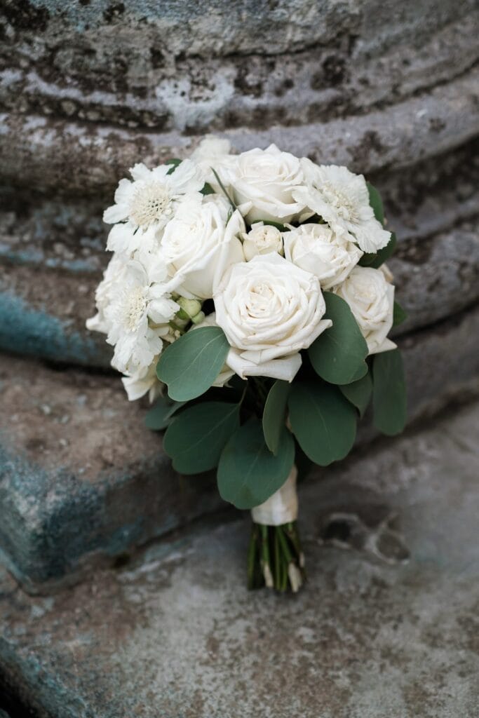 Hochzeitskrone Ein Strauß weißer Rosen und Grünpflanzen ruht auf einer Steinoberfläche und fängt die Essenz natürlicher Hochzeitsfotos ein.