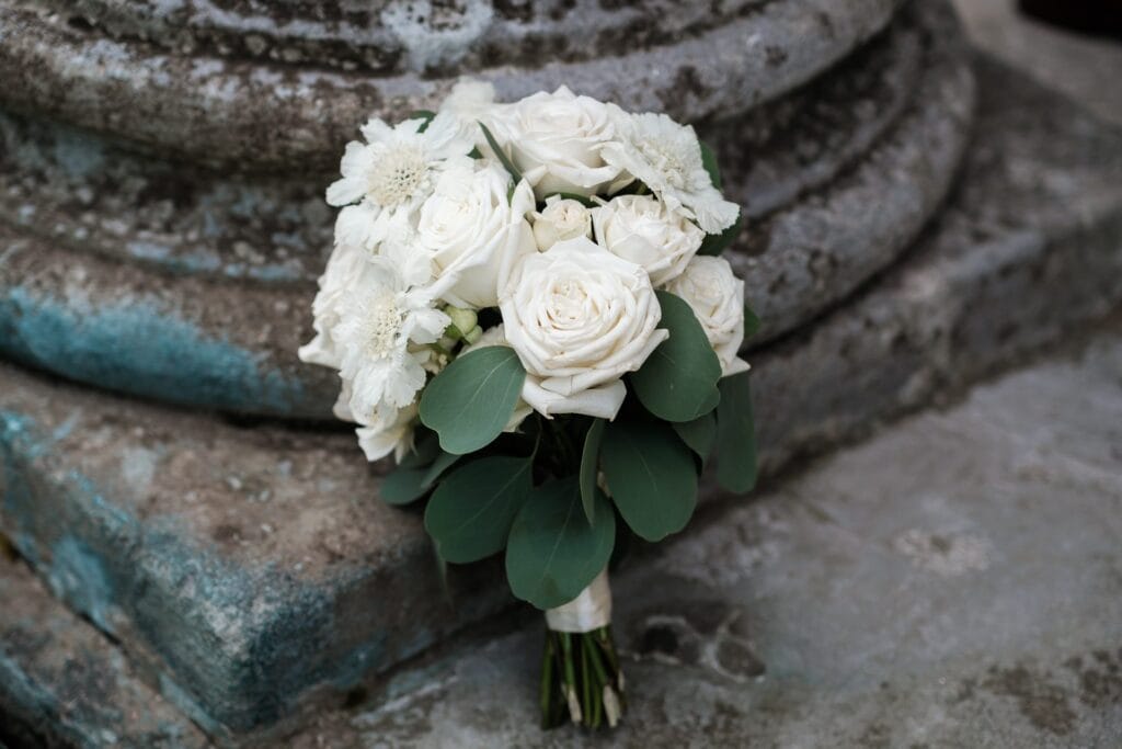 Hochzeitskrone Ein Strauß aus weißen Rosen und Grünpflanzen ruht auf einer Steinstruktur und fängt echte Momente ein, die an natürliche Hochzeitsfotos eines Hochzeitsfotografen in Wien und Niederösterreich erinnern.