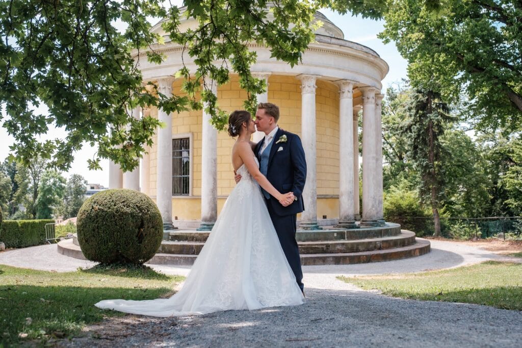 Hochzeitskrone Ein Paar im Hochzeitskleid küsst sich vor einem gelben, von Säulen umgebenen Pavillon, wunderschön festgehalten in authentischen Hochzeitsfotos, die ihre echte Liebe und Freude widerspiegeln.