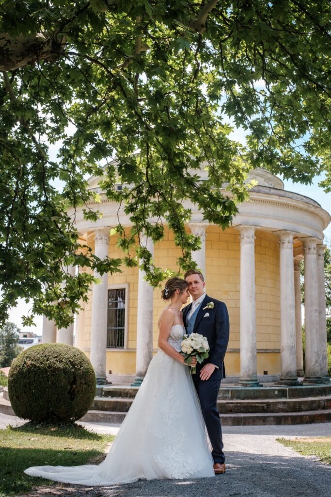 Hochzeitskrone Braut und Bräutigam, aufgenommen von einem Hochzeitsfotografen in Wien und Niederösterreich, posieren vor einem gelben Pavillon mit Säulen. Sie hält einen Blumenstrauß in der Hand, während sie an einem sonnigen Tag unter Ästen stehen und verkörpert die Essenz natürlicher Hochzeitsfotos.