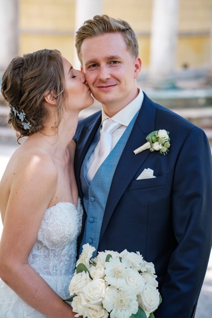 Hochzeitskrone Die Braut im weißen Kleid küsst den Bräutigam im blauen Anzug auf die Wange, während er einen Strauß weißer Blumen hält. Ihr freudiger Moment fängt die Essenz natürlicher Hochzeitsfotos ein und unterstreicht die Schönheit der Liebe, festgehalten von einem erfahrenen Hochzeitsfotografen in Wien und Niederösterreich.