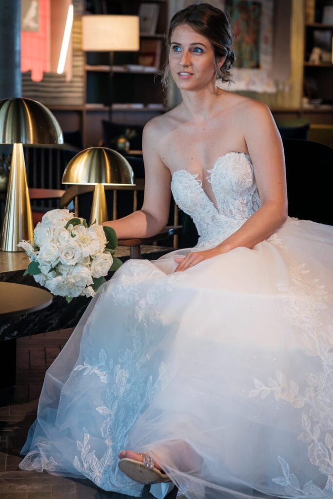 Hochzeitskrone Eine Braut in einem Spitzenbrautkleid hält einen Strauß weißer Rosen in der Hand, sitzt im Innenbereich neben goldenen Lampen und dunklen Stühlen und fängt echte Momente mit authentischen Hochzeitsfotos ein, die die Emotionen des Tages wirklich widerspiegeln.