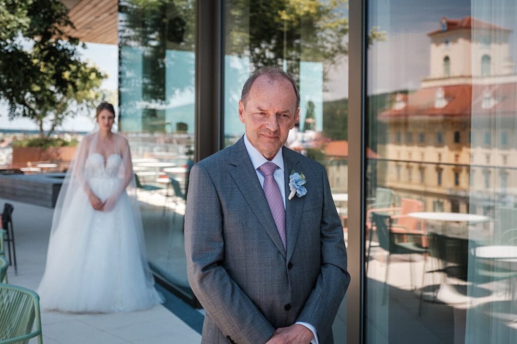 Hochzeitskrone Ein Mann im Anzug steht scharf auf der Terrasse und macht eines dieser authentischen Hochzeitsfotos, während im Hintergrund eine Frau im Brautkleid sanft unscharf ist.