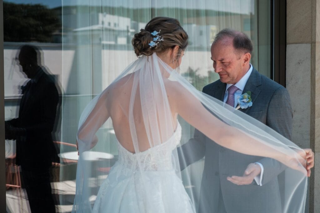 Hochzeitskrone Eine Braut in weißem Kleid und Schleier steht mit einem älteren Mann im Anzug im Haus. Der Mann hält den Schleier, während sie einander gegenüberstehen, ihr Bild spiegelt sich in einem Glasfenster – authentische Hochzeitsfotos, die echte Momente einfangen.