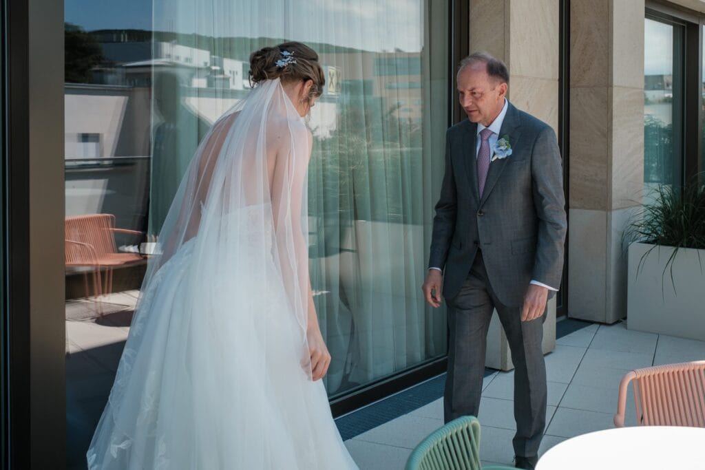 Hochzeitskrone Eine Braut in einem weißen Kleid und Schleier steht draußen auf einer Terrasse, einem älteren Mann in einem grauen Anzug mit einer Boutonniere gegenüber. In der Nähe sind Stühle ordentlich neben einer Glastür angeordnet – eine perfekte Szene für natürliche Hochzeitsfotos, aufgenommen von einem erfahrenen Hochzeitsfotografen in Wien und Niederösterreich.