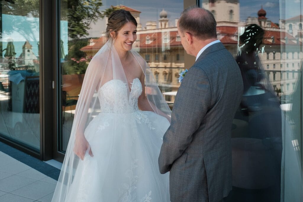 Hochzeitskrone Eine Braut in weißem Brautkleid und Schleier lächelt einen älteren Mann im Anzug auf einer Dachterrasse mit Blick auf die Stadt an und fängt echte Freude ein. Diese authentischen Hochzeitsfotos sind wertvolle Erinnerungen, die von einem erfahrenen Hochzeitsfotografen in Wien und Niederösterreich fotografiert wurden.