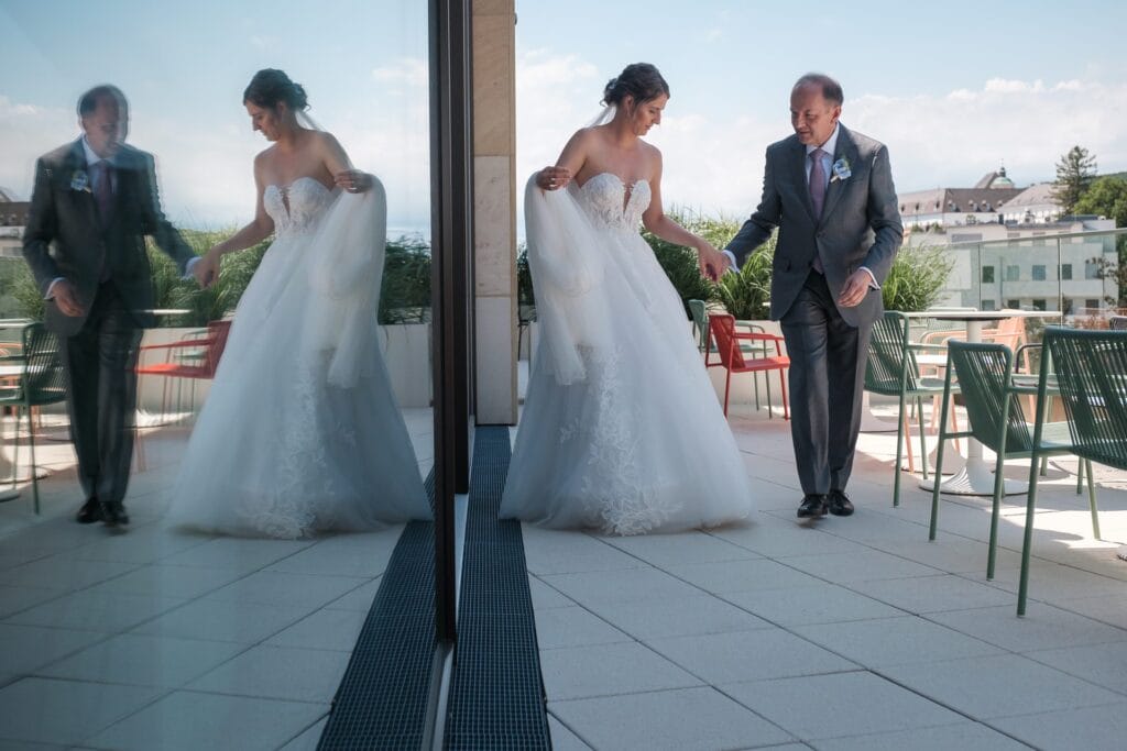 Hochzeitskrone Braut und älterer Mann, möglicherweise ihr Vater, gehen Hand in Hand über eine Terrasse mit Spiegelungen im Glas, umgeben von grünen Stühlen und Tischen. Perfekt für natürliche Hochzeitsfotos von einem Hochzeitsfotografen in Wien und Niederösterreich.