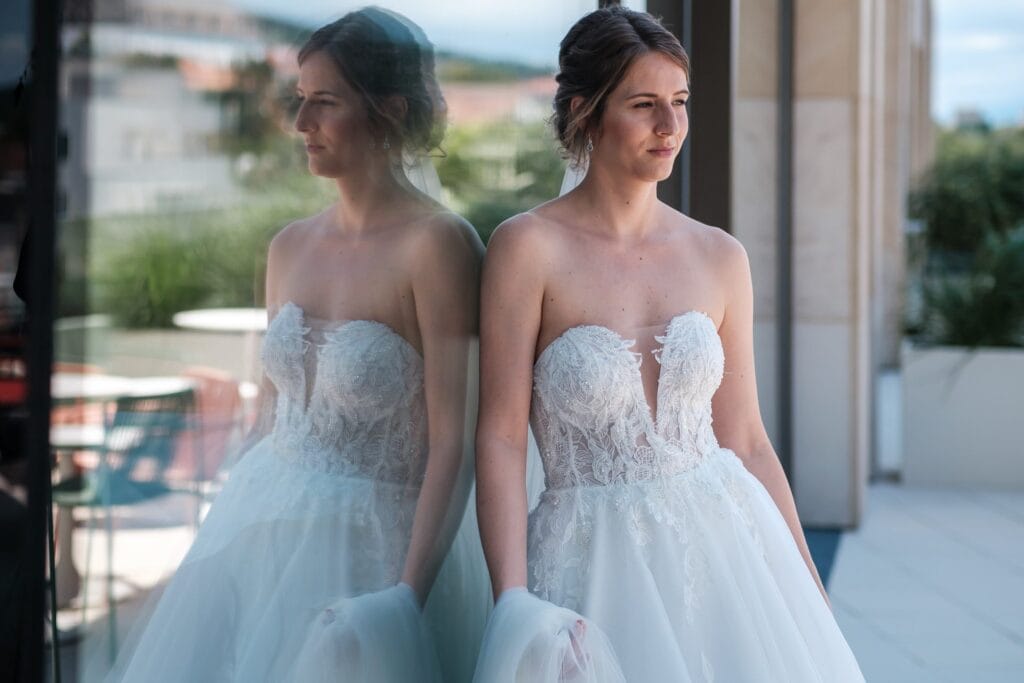 Hochzeitskrone Eine Frau in einem trägerlosen weißen Spitzenbrautkleid steht an einer reflektierenden Glaswand und blickt in die Ferne. Diese natürlichen Hochzeitsfotos fangen ihre heitere Schönheit und die zeitlose Eleganz ihres besonderen Tages perfekt ein.