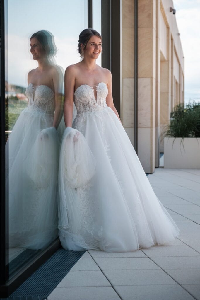 Hochzeitskrone Eine Braut in einem trägerlosen Spitzenbrautkleid steht mit ihrem Schleier vor einer reflektierenden Glaswand auf einer Terrasse und fängt echte Momente ein.