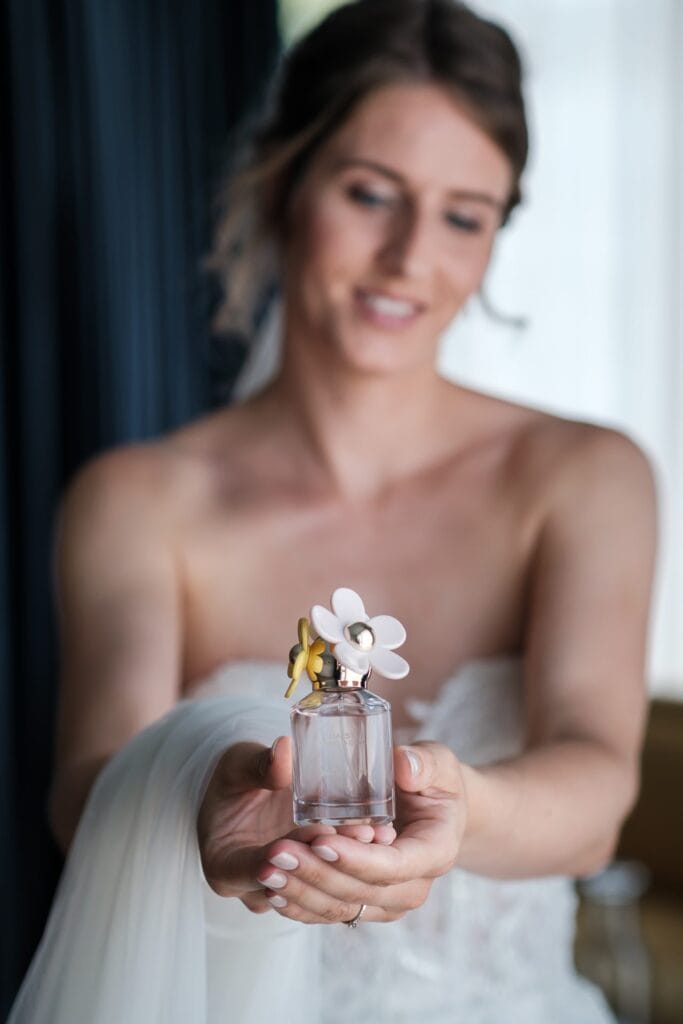 Hochzeitskrone Eine Frau im weißen Kleid hält einen Parfümflakon mit blumenförmiger Verzierung obendrauf, der mit seinem zarten Charme „echte Momente“ einfängt.