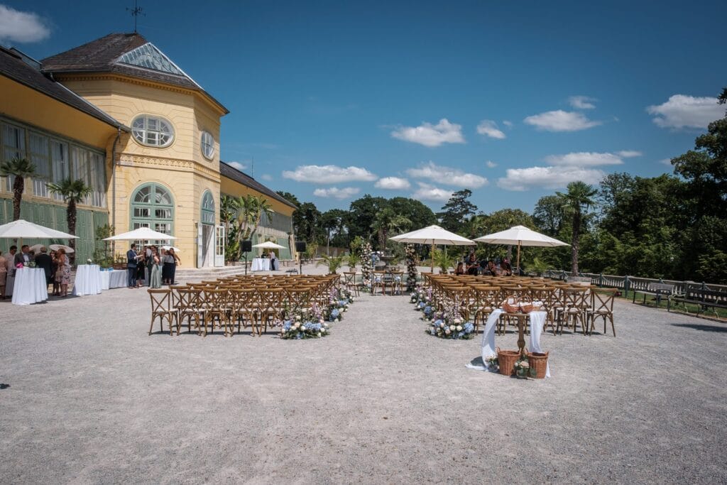 Hochzeitskrone Die Trauungszeremonie im Freien umfasst Holzstühle in Reihen, Blumendekorationen und weiße Sonnenschirme unter der Sonne, alles vor der Kulisse eines bezaubernden gelben Gebäudes. Diese malerische Kulisse ist perfekt für authentische Hochzeitsfotos, die echte Momente zelebrieren.