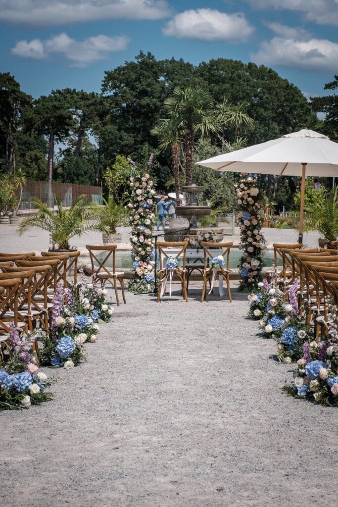 Hochzeitskrone Eine Hochzeitsdekoration im Freien mit Holzstühlen entlang eines mit Blumen gesäumten Ganges fängt echte Momente ein. Am Ende erwarten Sie zwei Blumensäulen und ein Tisch, eingerahmt von Bäumen und einem Brunnen im Hintergrund – perfekt für natürliche Hochzeitsfotos.