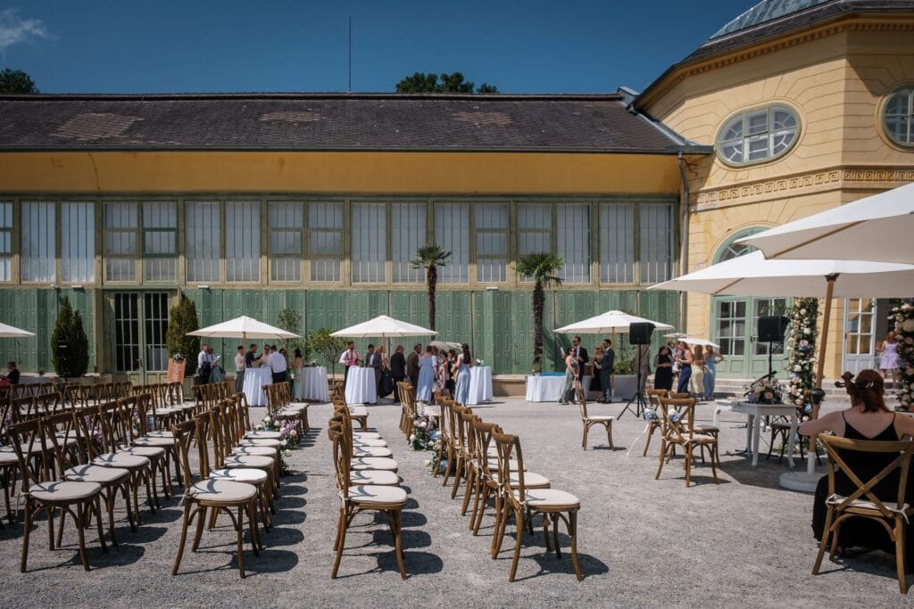 Hochzeitskrone Hochzeitsort im Freien mit Reihen leerer Holzstühle vor einem gelben Gebäude, perfekt für natürliche Hochzeitsfotos. Gäste mischen sich an Tischen mit Sonnenschirmen im Hintergrund unter einem klaren blauen Himmel und bieten eine ideale Kulisse für jeden Hochzeitsfotografen in Wien und Niederösterreich.