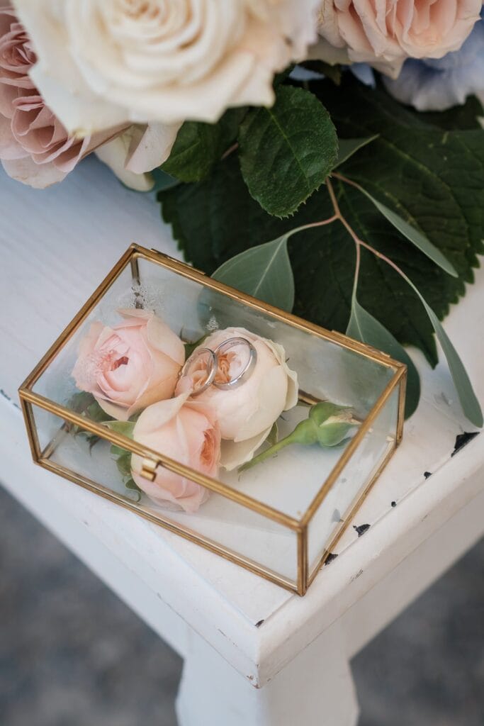 Hochzeitskrone Eine Glasbox mit Goldbesatz, die echte Momente einfängt, enthält zwei Ringe auf rosa Rosen. Es ruht elegant auf einer weißen Oberfläche neben einem Strauß Rosen und Grünpflanzen, perfekt für alle, die authentische Hochzeitsfotos von einem Hochzeitsfotografen in Wien und Niederösterreich suchen.