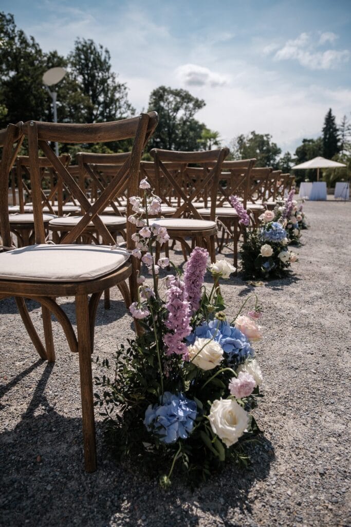 Hochzeitskrone Bei einer sonnendurchfluteten Hochzeitszeremonie im Freien stehen Holzstühle in Reihen und der Gang ist mit rosa, blauen und weißen Blumen geschmückt. Es ist eine malerische Kulisse, perfekt für authentische Hochzeitsfotos, die echte Momente der Freude und Liebe festhalten.