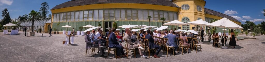 Hochzeitskrone Veranstaltung im Freien mit Menschen, die unter weißen Sonnenschirmen in der Nähe eines großen gelben Gebäudes mit einem Glasanbau sitzen, perfekt für die Aufnahme authentischer Hochzeitsfotos.