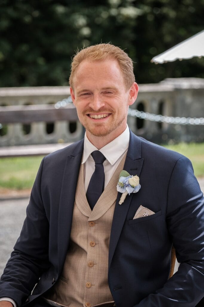 Hochzeitskrone Ein Mann in einem marineblauen Anzug mit beiger Weste und Krawatte sitzt lächelnd im Freien, hat kurze Haare und fängt echte Momente ein. Die verschwommenen Bäume und Steinstrukturen hinter ihm bilden eine ideale Kulisse, die Hochzeitsfotografen in Wien und Niederösterreich häufig sehen.