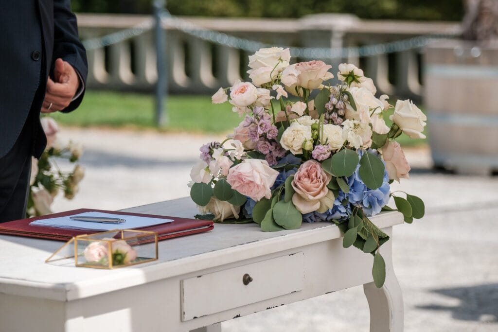 Hochzeitskrone Ein weißer Tisch mit einem Blumenarrangement, einem geschlossenen roten Buch und einer kleinen Schachtel steht daneben, während eine Person im dunklen Anzug bereitsteht, um echte Momente festzuhalten.