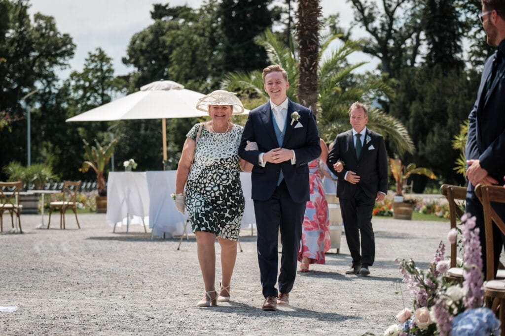 Hochzeitskrone Ein Mann im Anzug begleitet eine Frau in einem geblümten Kleid und Hut einen Gang im Freien entlang und fängt dabei echte Momente der Freude ein. Zwei weitere folgen ihm.