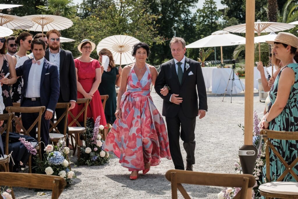 Hochzeitskrone Eine Frau im Blumenkleid geht Arm in Arm mit einem Mann im Anzug einen Gang im Freien entlang, umgeben von Gästen, die auf Holzstühlen sitzen. Es ist ein Bilderbuchtag, gefüllt mit authentischen Hochzeitsfotos, die jeden echten Moment dieser sonnigen Feier festhalten.