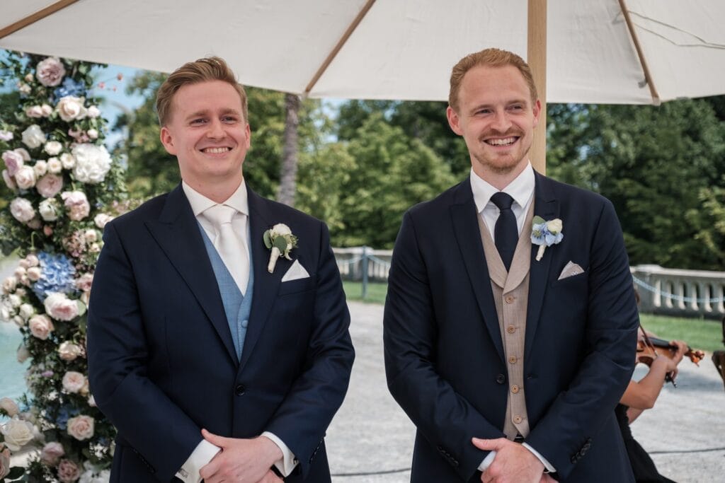 Hochzeitskrone Zwei Männer in Anzügen stehen lächelnd unter einem Regenschirm bei einer Veranstaltung im Freien, umgeben von Blumendekorationen und einem Musiker im Hintergrund. Diese authentischen Hochzeitsfotos fangen echte Momente ein und zeigen die freudige Stimmung des Anlasses.