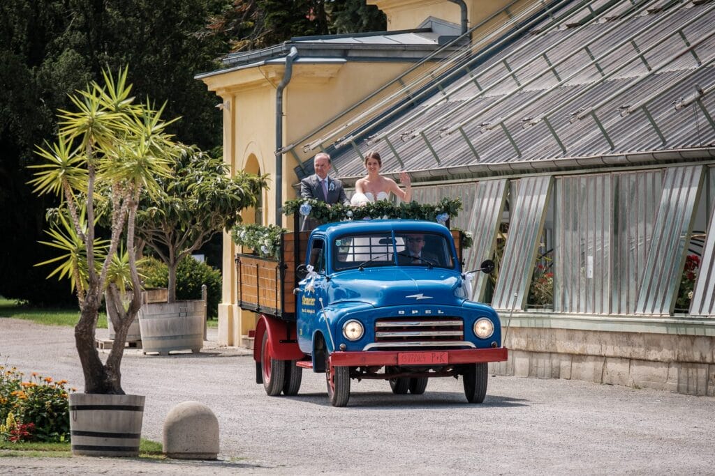 Hochzeitskrone Braut und Bräutigam stehen auf der Ladefläche eines mit Grünpflanzen geschmückten blauen Opel-Oldtimers, der vor einem Gebäude im Gewächshausstil geparkt ist. Fotografiert wurde das Bild von einem Hochzeitsfotografen in Wien und Niederösterreich, der auf authentische Hochzeitsfotos spezialisiert ist, die echte Momente hervorheben.