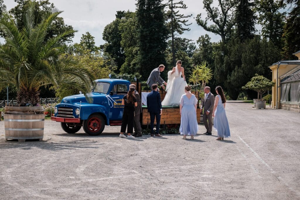 Hochzeitskrone Eine Braut in einem weißen Kleid steht auf der Ladefläche eines blauen Oldtimer-Trucks, umgeben von Menschen, und macht authentische Hochzeitsfotos. Die Außenkulisse, eingerahmt von Bäumen und einem Gebäude, verleiht der Szene Charme und echte Emotionen.