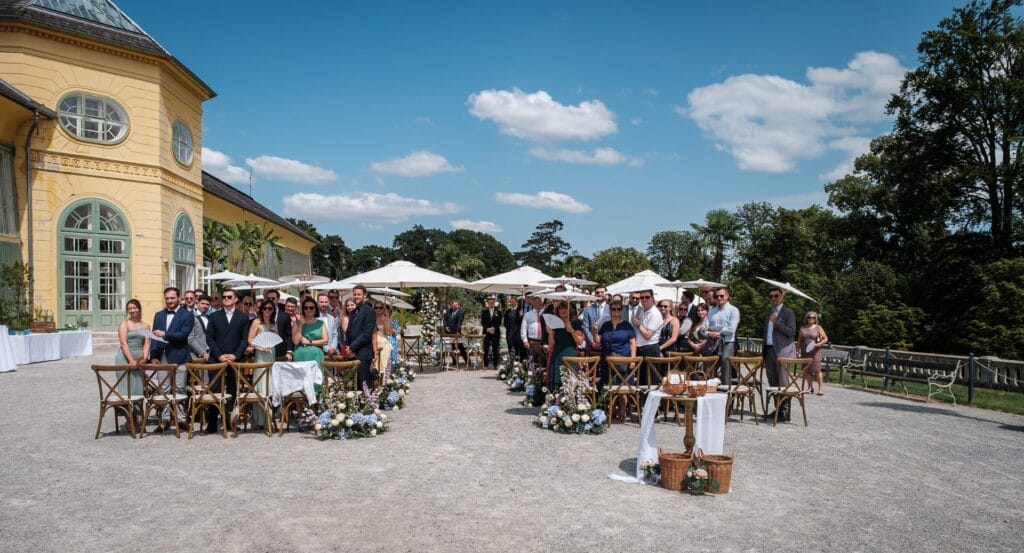 Hochzeitskrone Hochzeitszeremonie im Freien mit Gästen auf Holzstühlen, umgeben von Blumenarrangements. Authentische Momente entfalten sich unter klarem Himmel neben einem gelben Gebäude. Perfekt für authentische Hochzeitsfotos, aufgenommen von einem talentierten Hochzeitsfotografen in Wien und Niederösterreich.