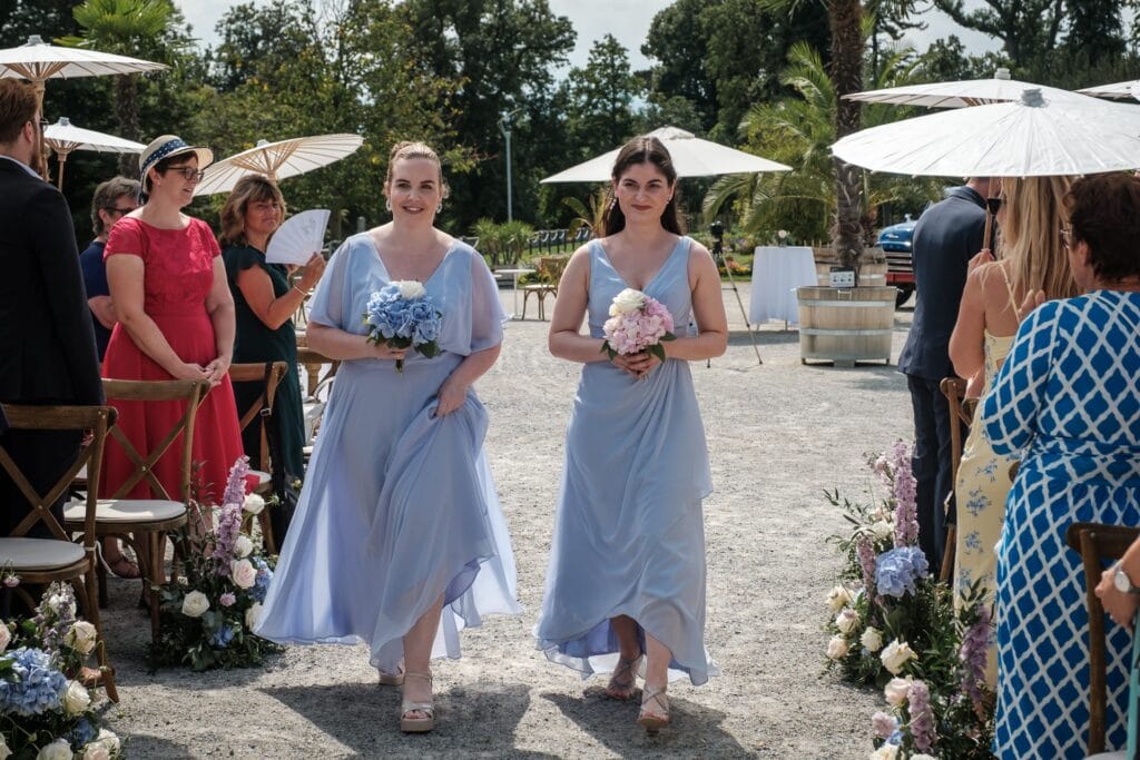 Hochzeitskrone Zwei Frauen in hellblauen Kleidern gehen einen Gang im Freien entlang, jede hält einen Blumenstrauß in der Hand. Gäste auf beiden Seiten sitzen unter weißen Sonnenschirmen und fangen echte Momente der Freude und des Zusammenseins ein.