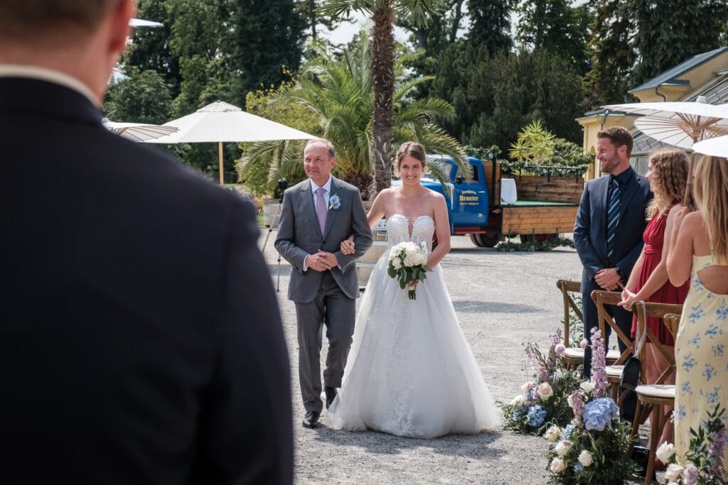 Hochzeitskrone Die Braut schreitet anmutig mit einem älteren Mann den Gang entlang und hält dabei einen Blumenstrauß in der Hand. Die Gäste sitzen rechts in dieser ruhigen Außenkulisse mit Bäumen und Sonnenschirmen. Halten Sie diese echten Momente mit authentischen Hochzeitsfotos von einem Hochzeitsfotografen in Wien und Niederösterreich fest.