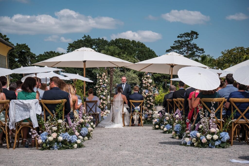 Hochzeitskrone Bei dieser bezaubernden Hochzeitszeremonie im Freien steht das Paar dem Geistlichen gegenüber, während die Gäste unter weißen Sonnenschirmen inmitten atemberaubender Blumenarrangements sitzen. Ein Hochzeitsfotograf in Wien und Niederösterreich hält jeden Moment fest und ist auf authentische Hochzeitsfotos spezialisiert, um diese wertvollen Erinnerungen zu bewahren.