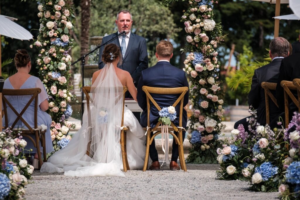 Hochzeitskrone Ein Paar sitzt während einer Trauungszeremonie im Freien einem Trauzeugen gegenüber, umgeben von Blumenarrangements in sanften Farben. Eingefangen von einem Hochzeitsfotografen in Wien und Niederösterreich, versprüht die Szene den Charme authentischer Hochzeitsfotos.