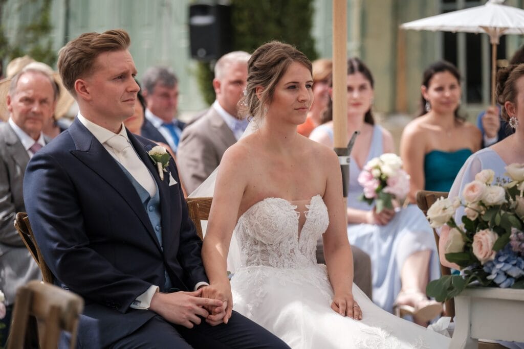 Hochzeitskrone Eine Braut und ein Bräutigam sitzen während einer Hochzeitszeremonie im Freien Händchen haltend zusammen und sind von Gästen umgeben. Dieser Moment wird wunderschön eingefangen von einem Hochzeitsfotografen in Wien und Niederösterreich, spezialisiert auf natürliche Hochzeitsfotos.