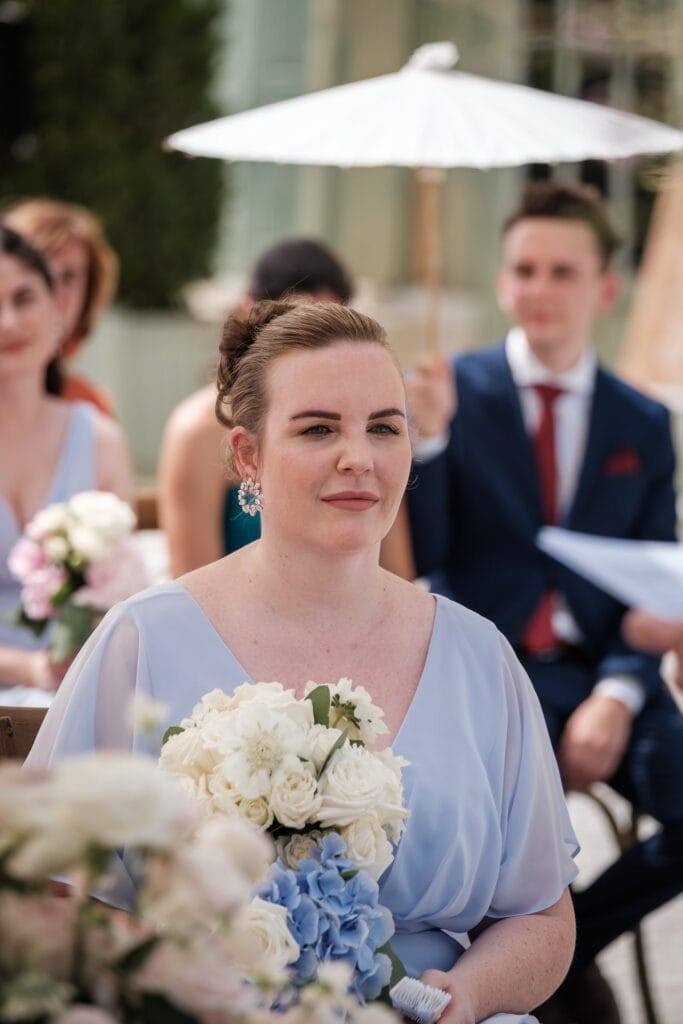 Hochzeitskrone Eine Frau in einem hellblauen Kleid hält einen Strauß weißer und blauer Blumen und fängt damit echte Momente ein. Sie ist im Freien, mit sitzenden Gästen und einer Person, die im Hintergrund einen weißen Sonnenschirm hält – perfekt für authentische Hochzeitsfotos von einem Hochzeitsfotografen in Wien und Niederösterreich.