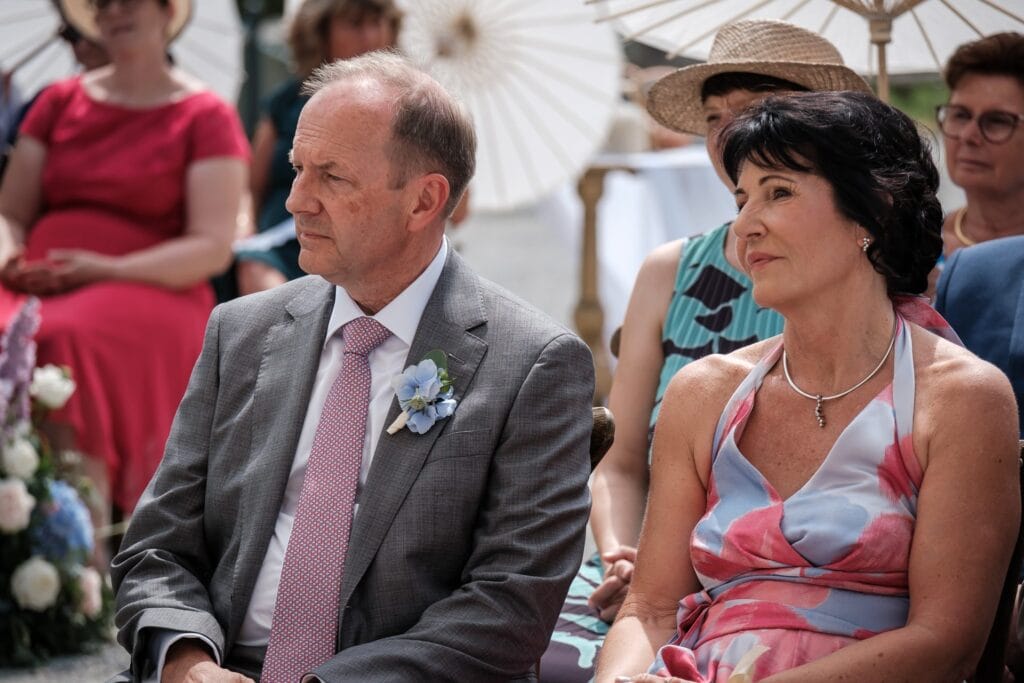 Hochzeitskrone Ein Mann im grauen Anzug mit einer Blumenboutonniere sitzt neben einer Frau im bunten Kleid, die einen Regenschirm hält. Umgeben von anderen genießen sie die echte Atmosphäre des Tages und fangen echte Momente voller natürlichem Charme und Freude ein, ganz wie authentische Hochzeitsfotos im Freien.