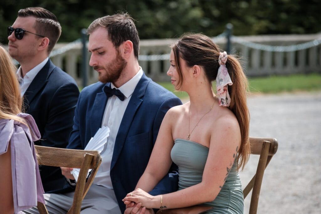 Hochzeitskrone Zwei Personen sitzen im Freien auf Holzstühlen; der Mann im blauen Anzug und mit Fliege hält Papiere, während die Frau im schulterfreien Kleid mit floralem Haarschmuck echte Momente verkörpert. Aufgenommen von einem Hochzeitsfotografen in Wien und Niederösterreich, sind dies wirklich natürliche Hochzeitsfotos.