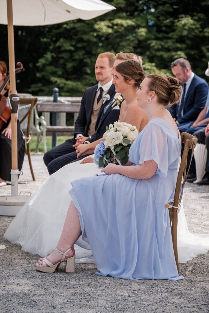 Hochzeitskrone Braut und Bräutigam sitzen im Freien mit Brautjungfern; eine Brautjungfer in Hellblau mit einem Strauß weißer Blumen fängt die Essenz authentischer Hochzeitsfotografie ein. Perfekt für alle, die echte Momente suchen, eine ideale Szene für einen Hochzeitsfotografen in Wien und Niederösterreich.