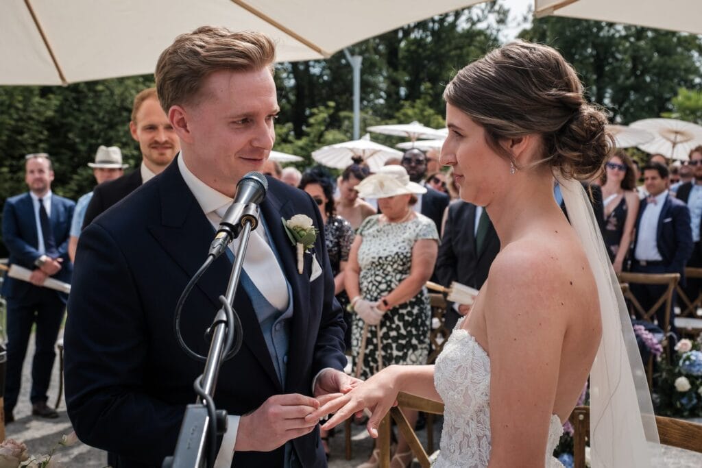 Hochzeitskrone Ein Paar tauscht während seiner Trauung im Freien die Ringe aus, perfekt festgehalten von einem Hochzeitsfotografen in Wien und Niederösterreich. Der Bräutigam hält die Hand der Braut an einem Mikrofonständer, unter Sonnenschirmen und bei sonnigem Wetter – echte Momente der Freude und Liebe.