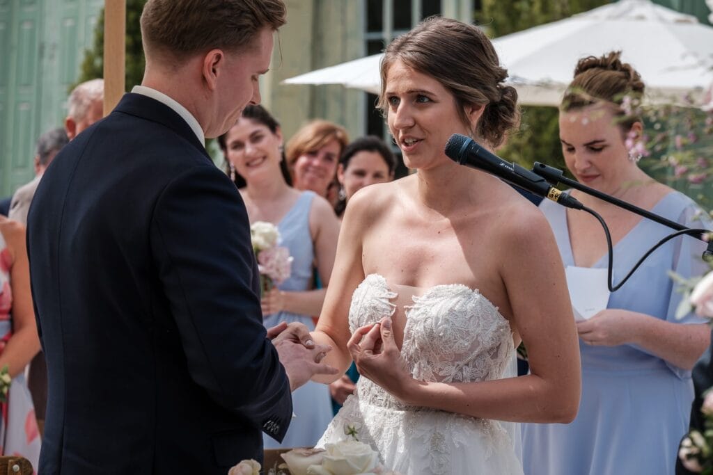 Hochzeitskrone Inmitten der ruhigen Außenkulisse tauschen Braut und Bräutigam herzliche Gelübde aus, die von einem Hochzeitsfotografen in Wien und Niederösterreich mühelos festgehalten werden. Die Gäste werden Zeuge dieser echten Momente, wobei das leise Summen eines Mikrofons die natürliche Atmosphäre verstärkt.