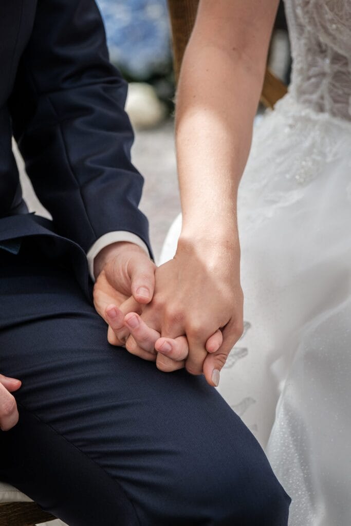 Hochzeitskrone Zwei Menschen in formeller Kleidung halten Händchen, sitzen eng beieinander und verkörpern mit ihrer echten Verbindung die Essenz natürlicher Hochzeitsfotos.