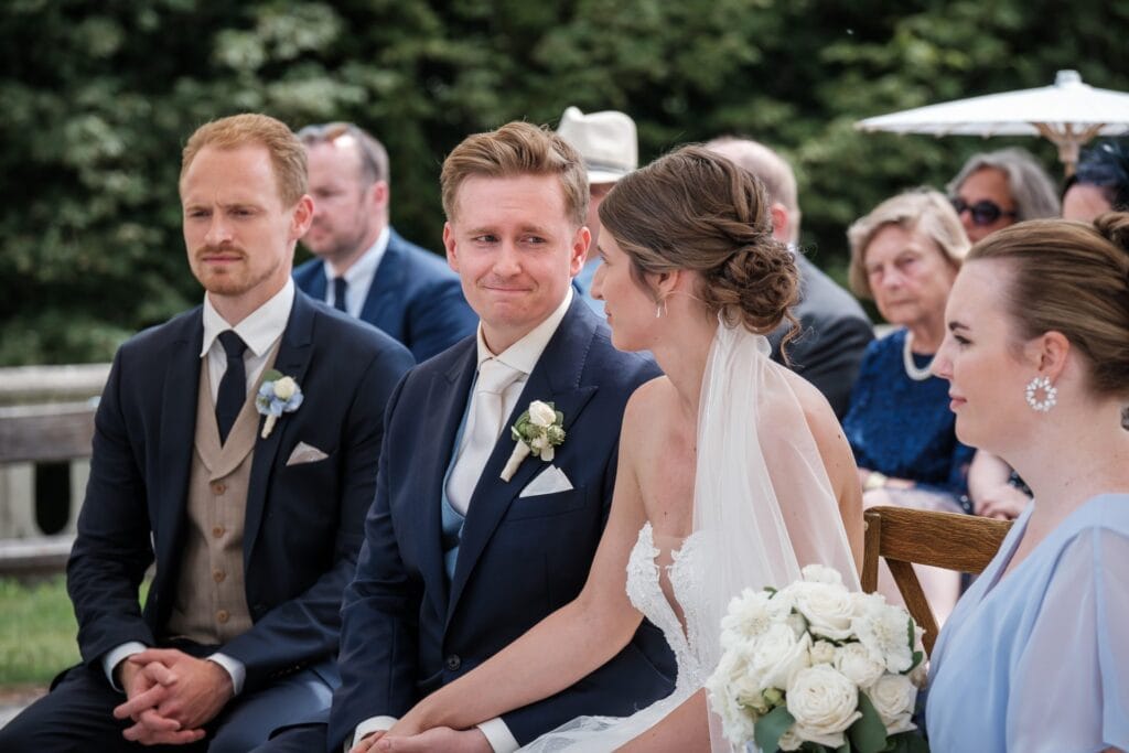 Hochzeitskrone Ein Paar in formeller Kleidung sitzt während einer Hochzeitszeremonie, umgeben von Gästen. Die Braut hält einen Strauß weißer Blumen und fängt echte Momente ein, die für authentische Hochzeitsfotos geeignet sind.